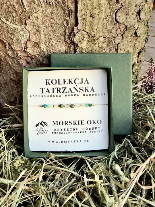 Bransoletka Morskie Oko z kolekcji Tatrzańskiej DM Tatra – ręcznie wykonana biżuteria z kryształem górskim, turmalinem, turkusem i apatytami, zaprezentowana w zielonym pudełku na tle kory drzewa i suszonej trawy.