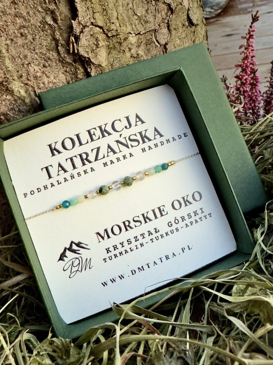 Bransoletka Morskie Oko z kolekcji Tatrzańskiej DM Tatra, wykonana z kryształu górskiego, turmalinu, turkusu i apatytu, prezentowana w zielonym pudełku na tle kory drzewa, wrzosów i suszonej trawy.