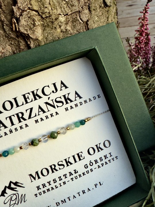 Zbliżenie bransoletki Morskie Oko z kolekcji Tatrzańskiej DM Tatra, wykonanej z kryształu górskiego, turmalinu, turkusu i apatytu, prezentowanej w zielonym pudełku na tle kory drzewa i roślin.