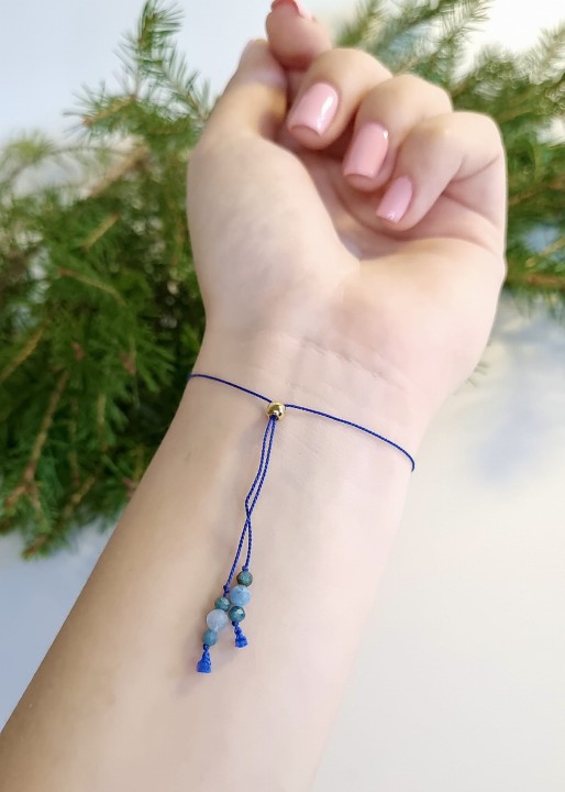 Bransoletka na niebieskim sznurku z drobnymi kamieniami w odcieniach turkusu i zieleni, prezentowana na nadgarstku osoby, na tle zielonych gałązek, w naturalnej i minimalistycznej aranżacji.