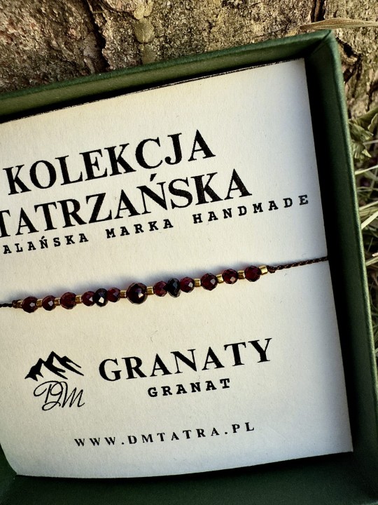 Zbliżenie bransoletki z naturalnych kamieni granatu z Kolekcji Tatrzańskiej DM Tatra, prezentowanej w zielonym pudełku na tle kory drzewa i suszonej trawy, ukazujące szczegóły ciemnoczerwonych fasetowanych koralików i złotych akcentów.