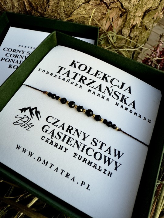 Zbliżenie bransoletki z czarnego turmalinu z Kolekcji Tatrzańskiej DM Tatra, prezentowanej w zielonym pudełku na tle kory drzewa i suszonej trawy; widoczne fasetowane czarne koraliki i złote akcenty w naturalnej, rustykalnej aranżacji.