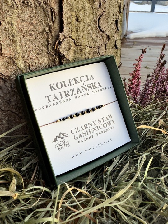 Bransoletka z czarnego turmalinu z Kolekcji Tatrzańskiej DM Tatra, prezentowana w zielonym pudełku na tle kory drzewa, suszonej trawy i wrzosów, w naturalnej zimowej aranżacji inspirowanej Tatrami.
