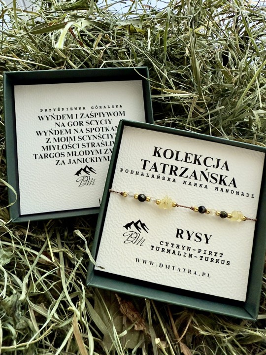 Bransoletka Rysy z cytrynu, pirytu, turmalinu i turkusu z Kolekcji Tatrzańskiej DM Tatra, prezentowana w zielonym pudełku na tle suszonej trawy; obok drugie pudełko z góralską przyśpiewką, tworzące naturalną, rustykalną aranżację.