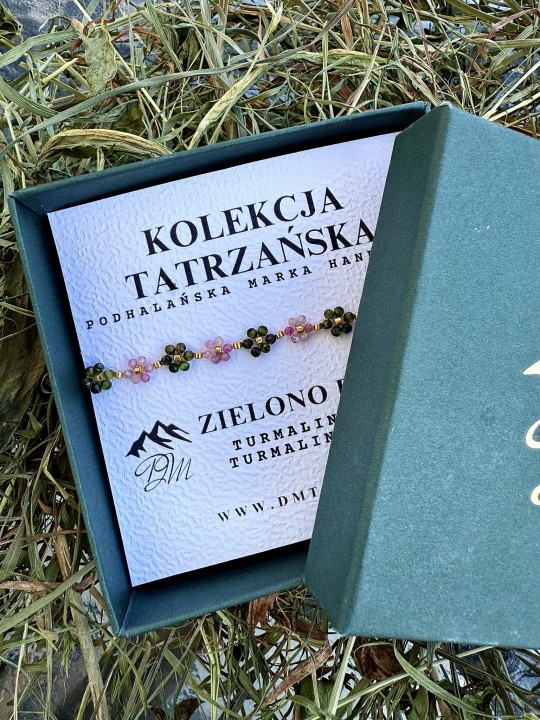 Bransoletka Zielono Polana z turmalinu różowego i zielonego, misternie pleciona na żyłce, z pozłacanymi koralikami ze stali chirurgicznej, prezentowana w zielonym pudełku DM Tatra na tle suszonej trawy.