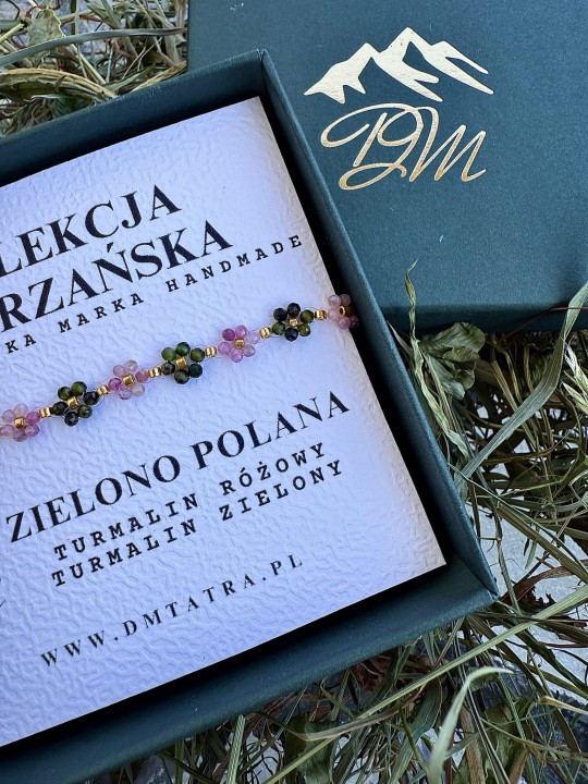 Zbliżenie bransoletki Zielono Polana z turmalinu różowego i zielonego plecionej na żyłce, z koralikami ze stali chirurgicznej, zaprezentowanej w pudełku DM Tatra na suszonej trawie