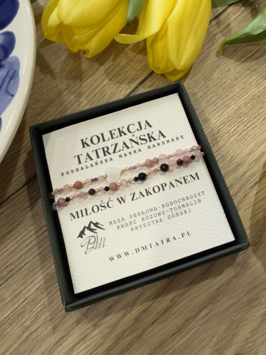 Bransoletka Miłość w Zakopanem z masy perłowej, rodochrozytu, kwarcu różowego, turmalinu i kryształu górskiego, prezentowana w zielonym pudełku Kolekcji Tatrzańskiej DM Tatra na drewnianym tle, obok żółtych tulipanów.